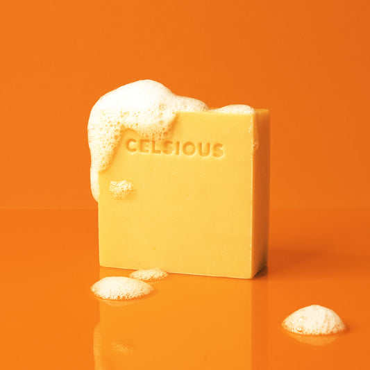 A block of yellow soap stamped with CELSIOUS photographed on vibrant orange with soft looking soap suds
