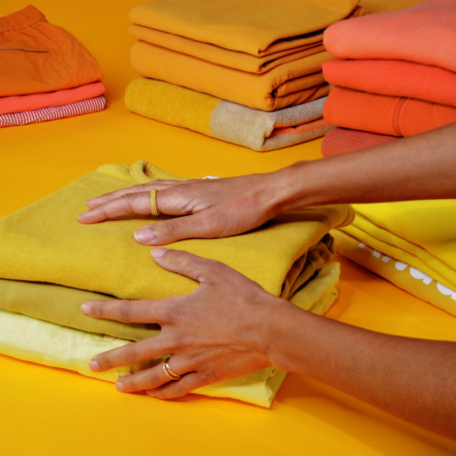 Two hands rest on a stack of neatly folded chartreuse shirts; neat stacks of orange and yellow clothes are visible nearby