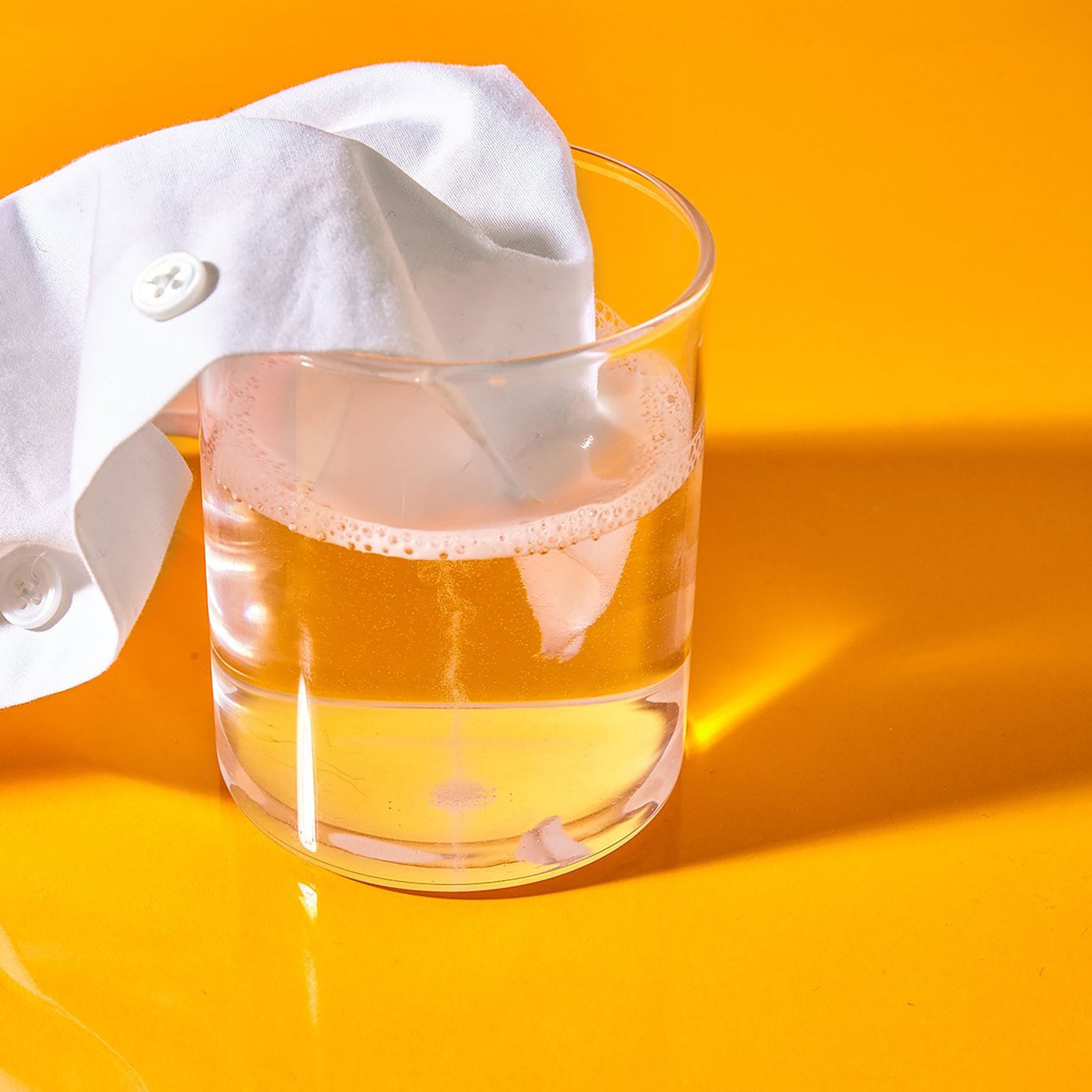 Supersalt oxygen brightener by Celsious dissolved in a clear glass of water and displayed with a white shirt against an orange backdrop