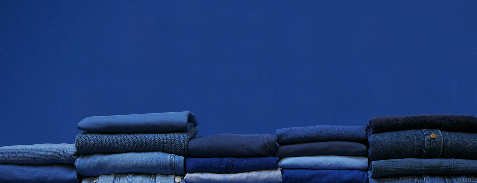 Neatly folded stacks of blue blue shirts, sweaters and denim jumpsuits in front of a blue backdrop