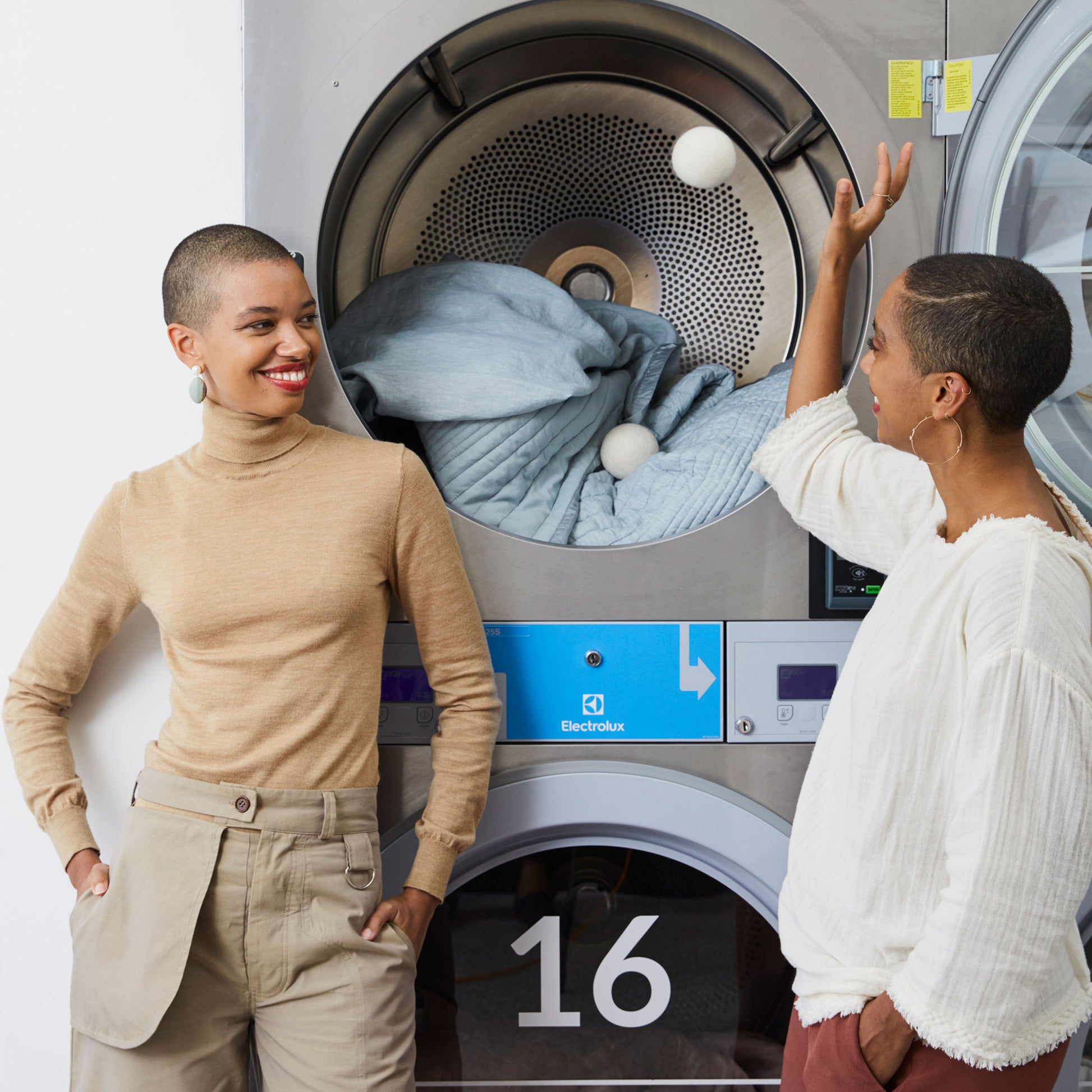 Theresa Williams smiles at her sister Corinna who is tossing a white wool dryer ball into a dryer filled with blue bed linens