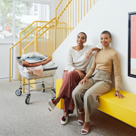 Corinna and Theresa Williams sit on a yellow bench in the bright entrance of their business; both wear outfits in earthy colors, and a laundry cart with fluffy linens is parked beside them