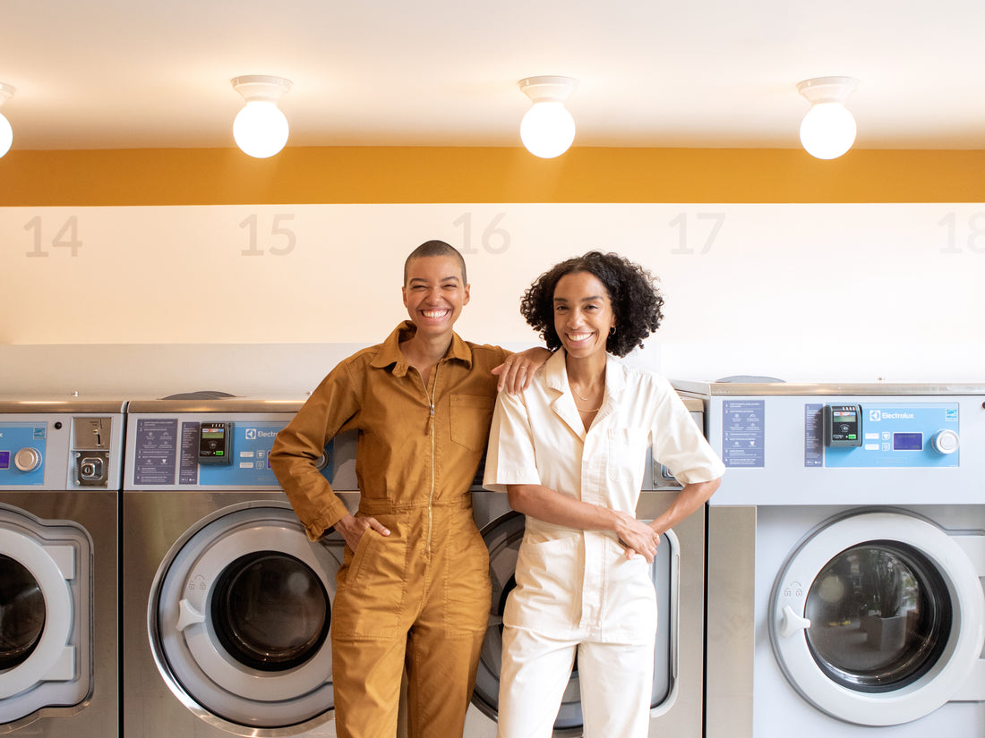 Corinna and Theresa Williams grin for the camera in front of dryers wearing stylish jumpsuits