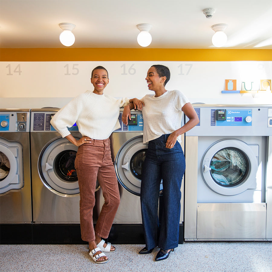 Two women in chic fall outfits laugh in a bright laundry space