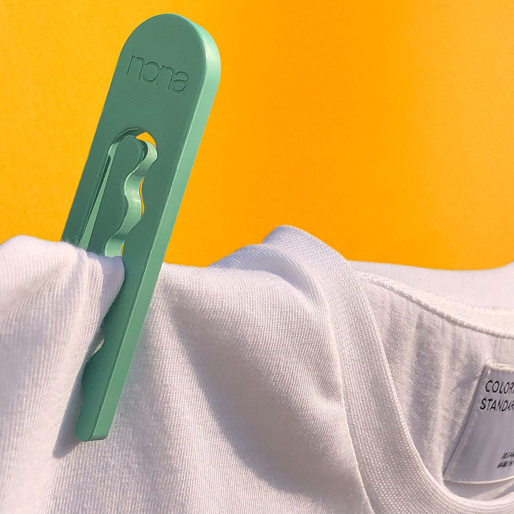 A green clothespin by NONA pins a white t-shirt in place on a clothesline