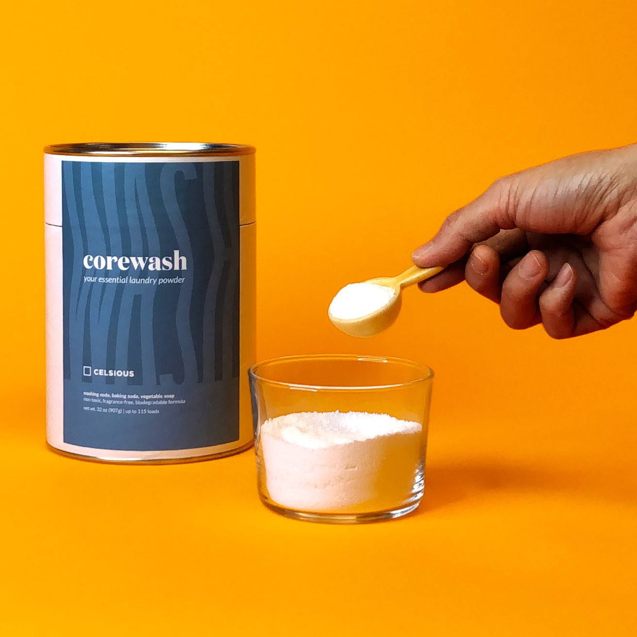 A canister of Corewash laundry powder by Celsious and a hand scooping laundry powder out of a glass with a tiny yellow scoop