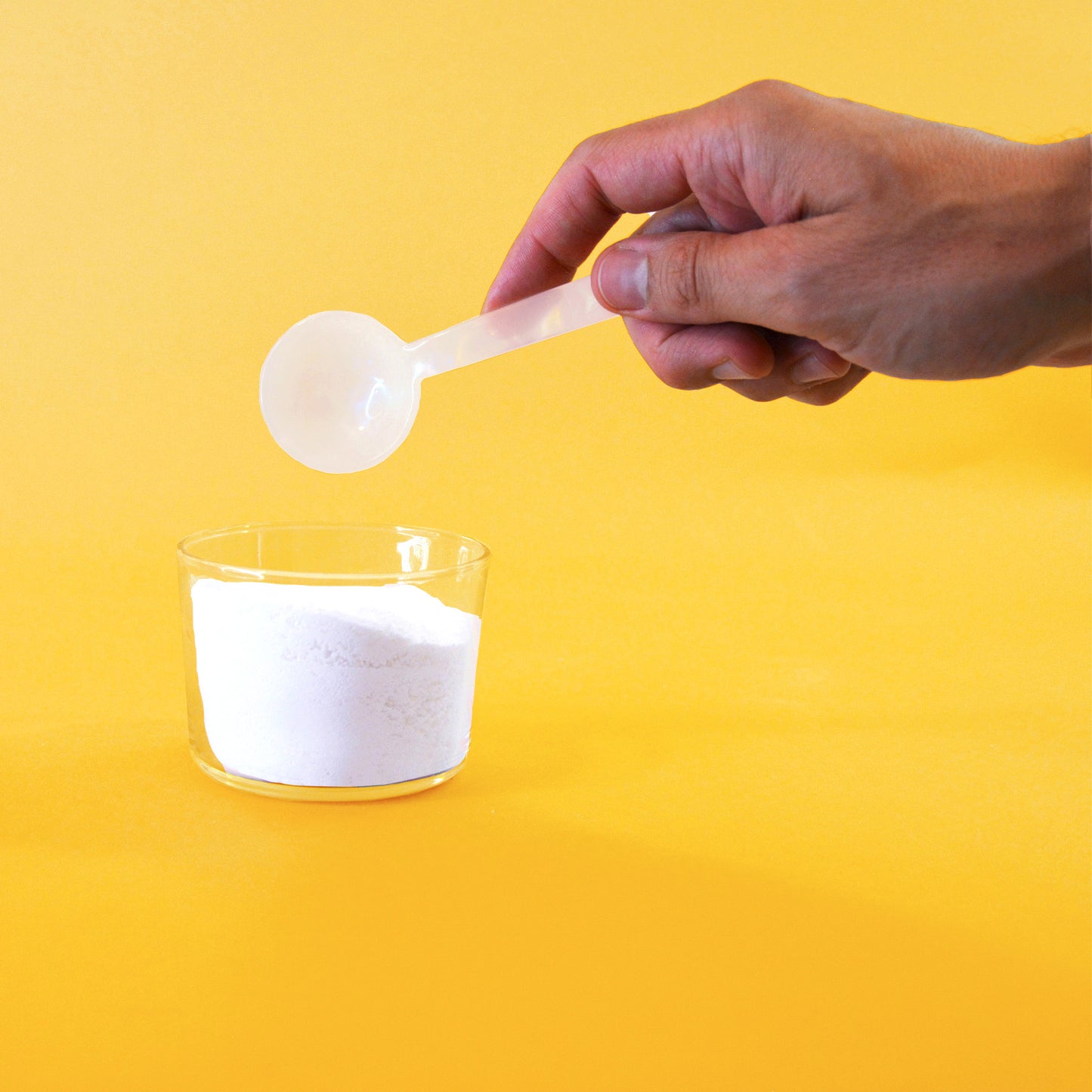 A hand holds an ivory cellulose spoon above a glass container filled with laundry powder.