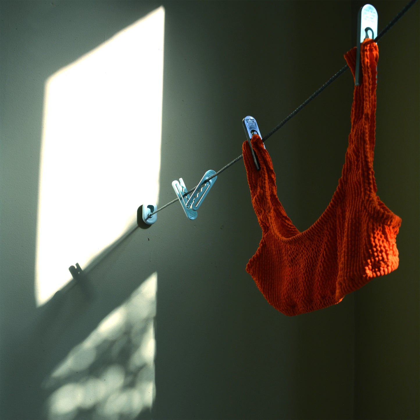 An orange swim top hangs from the LOOP clothesline held up by green NONA clothespins in the afternoon light