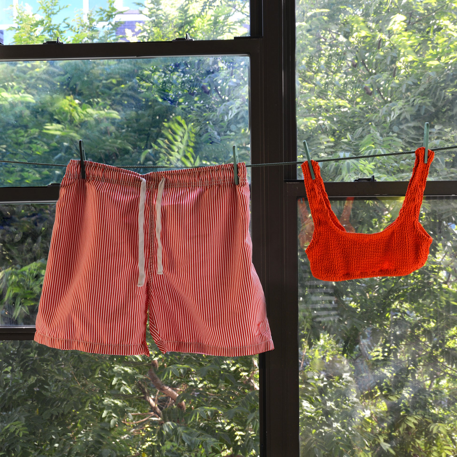 An orange swim top and striped swim trunks hang from a clothesline with clothespins; the background is leady