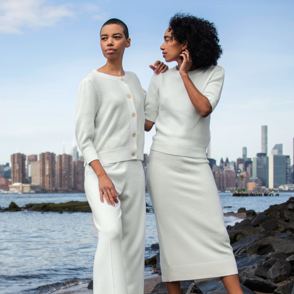Two models wearing matching off-white knit outfits stand on a rocky waterfront with a city skyline in the background. One model wears a cropped cardigan and wide-leg pants set, and the other wears a short-sleeve top and midi skirt set.