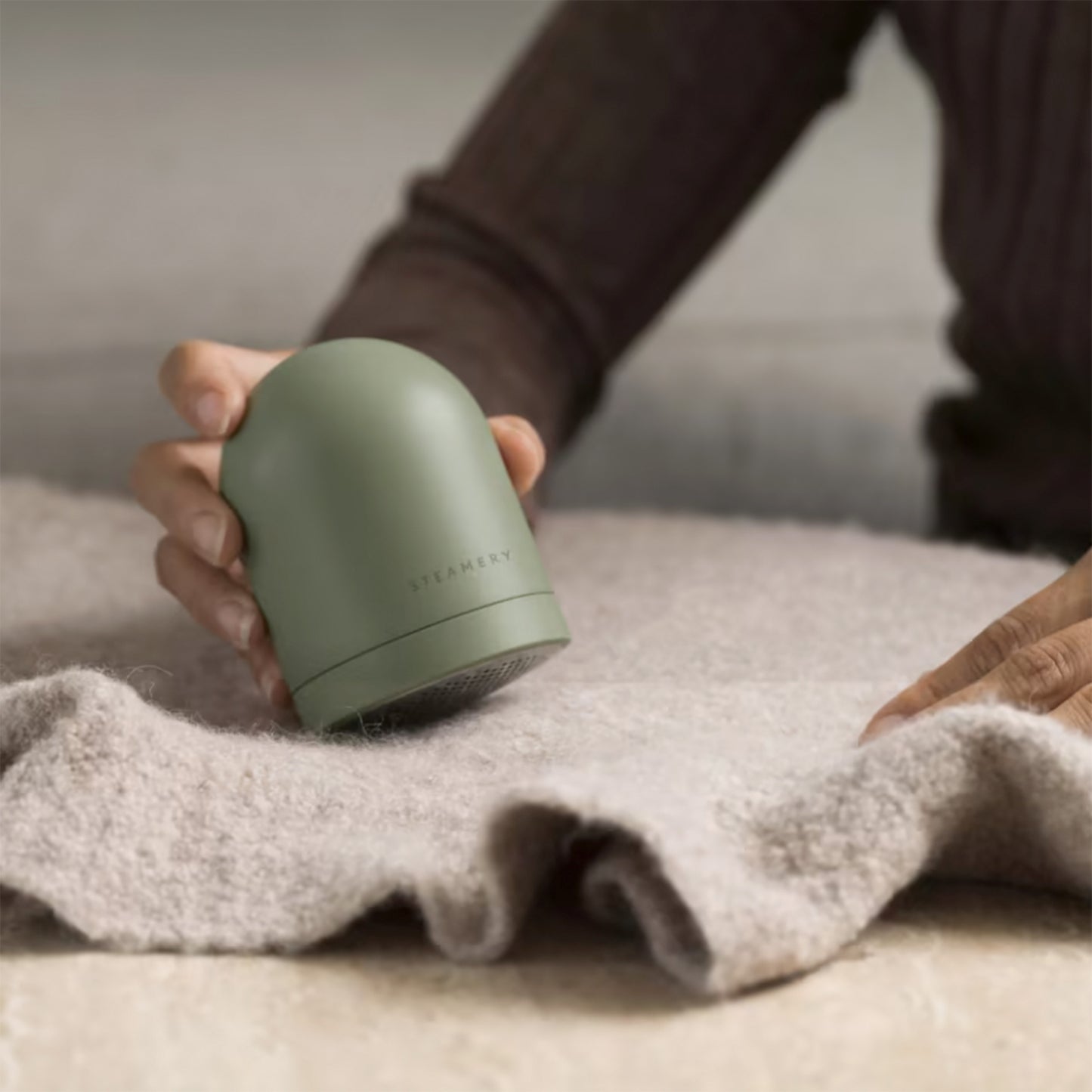A hand holds a sage "Pilo 2" fabric shaver up to a sweater