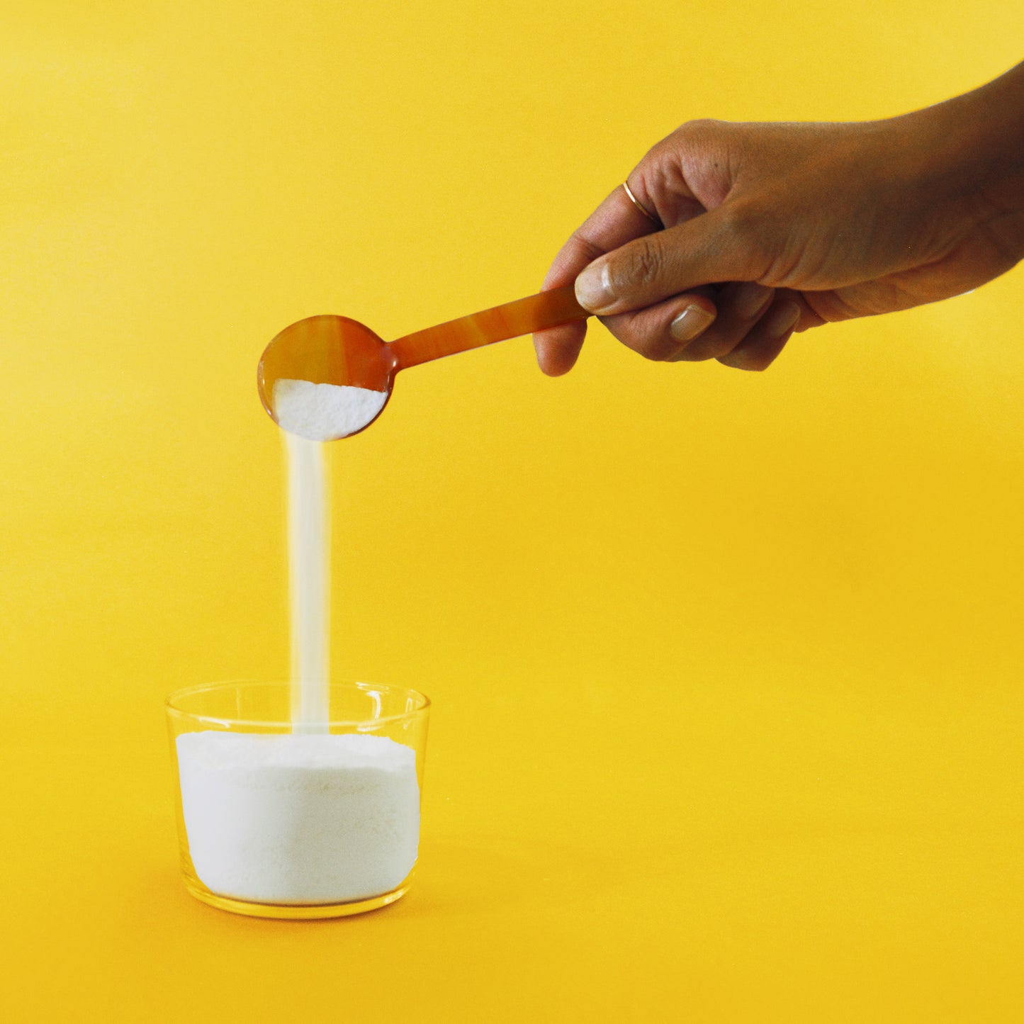 A hand tips a spoonful of laundry powder into a glass container below.