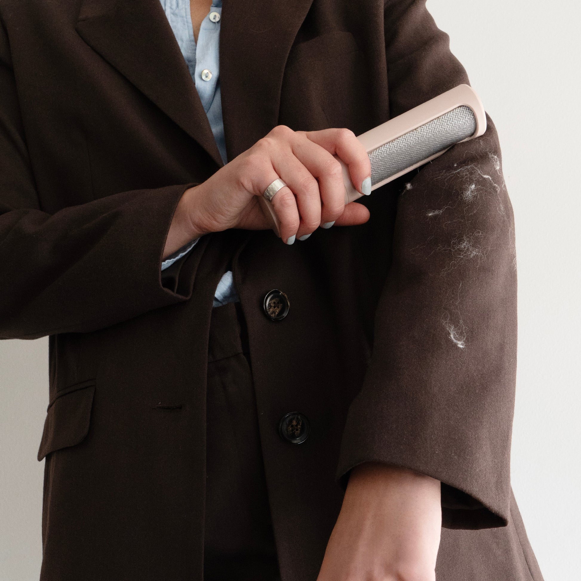 A woman brushes lint off her brown peacoat with a rosy pink reusable lint roller