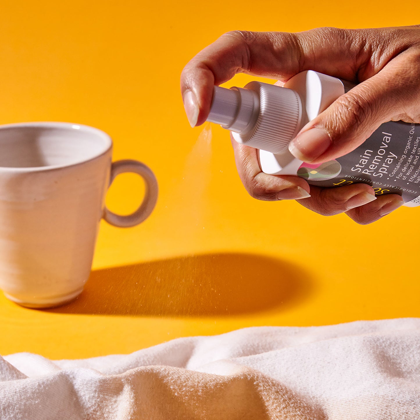 A hand sprays a white spray bottle of Sonett eco-friendly stain remover onto fabric stained with coffee; a mug is visible in the background