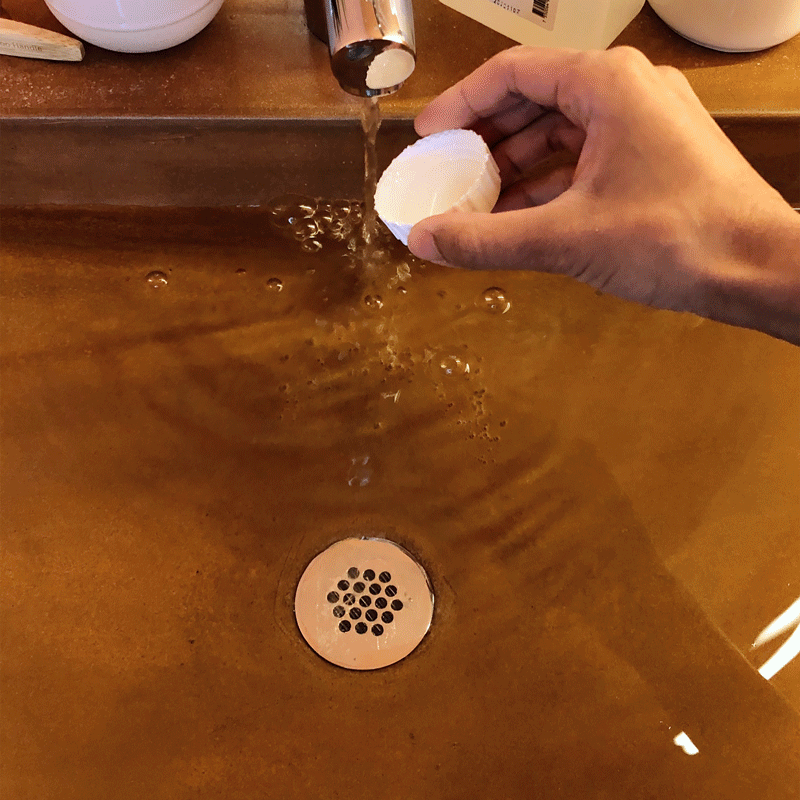 A gif of a hand pouring a cap full of delicate detergent into a clay sink, followed by a rose gold silk blouse submerged into the sudsy water by the same hand
