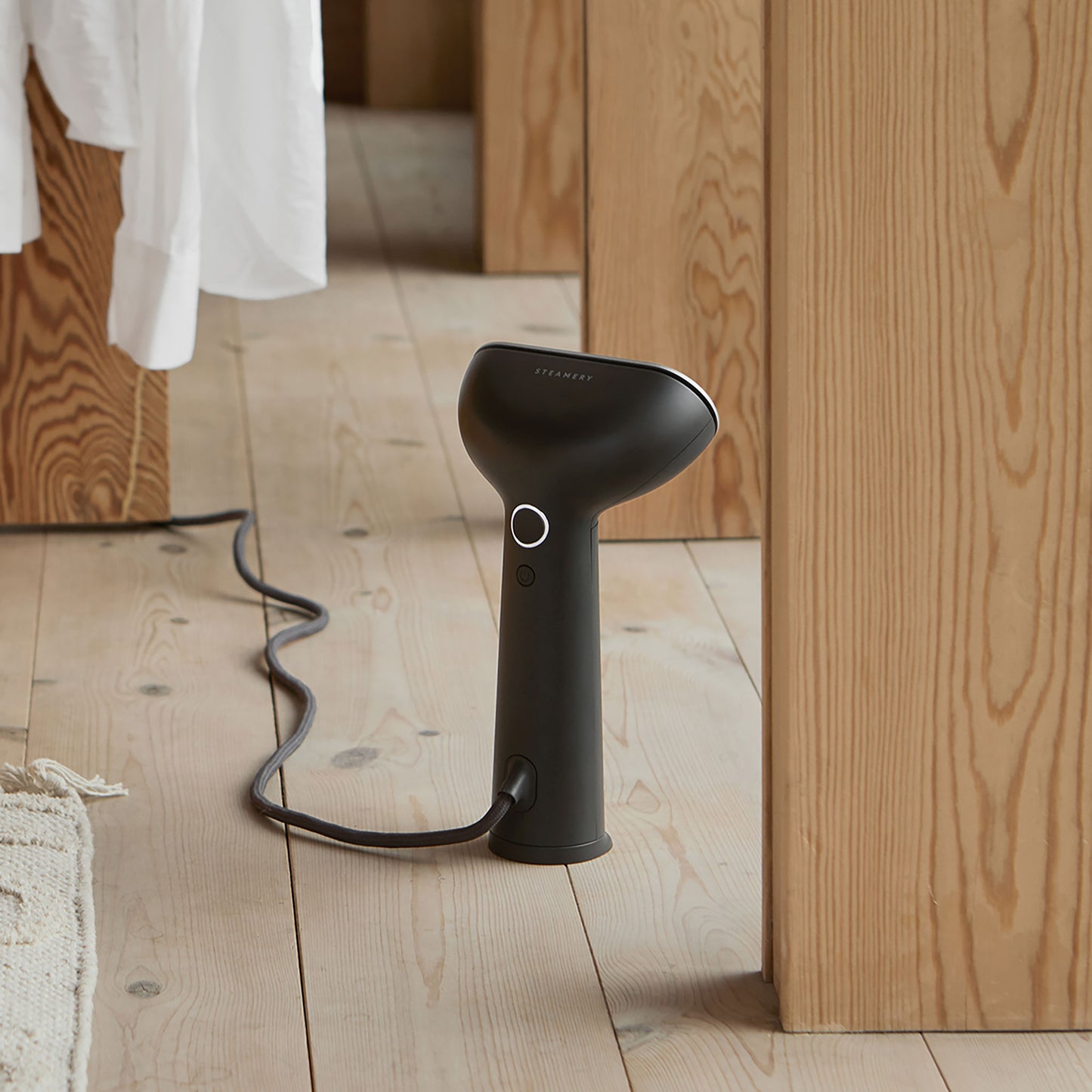 A sleek, black handheld device that functions as both a steamer and an iron, standing in a wooden cottage with the cord trailing behind it. A white linen shirt hangs in the background.