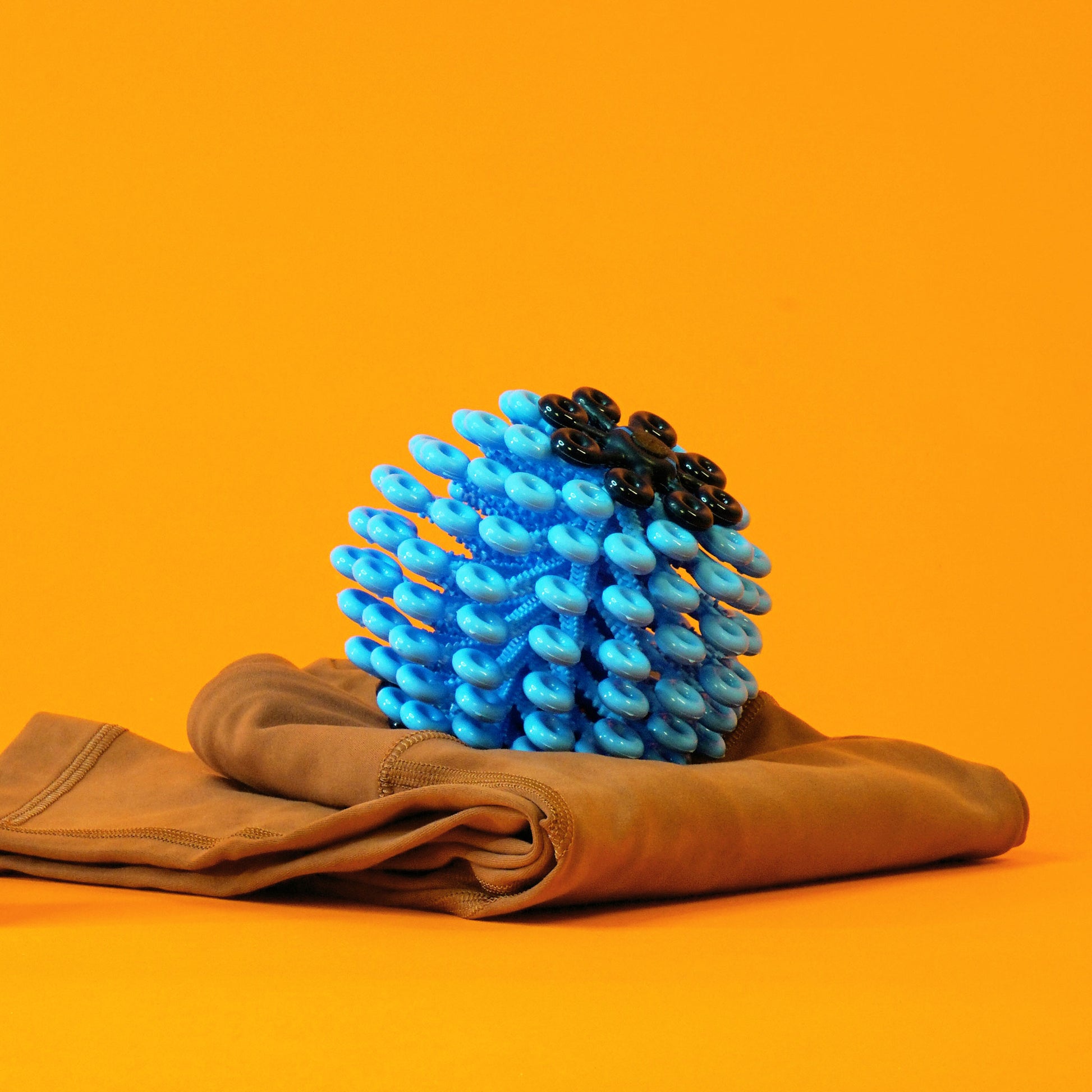 A blue laundry ball covered with numerous loops that is used to catch microfibers, displayed atop a folded pair of yoga leggings in front of an orange backdrop