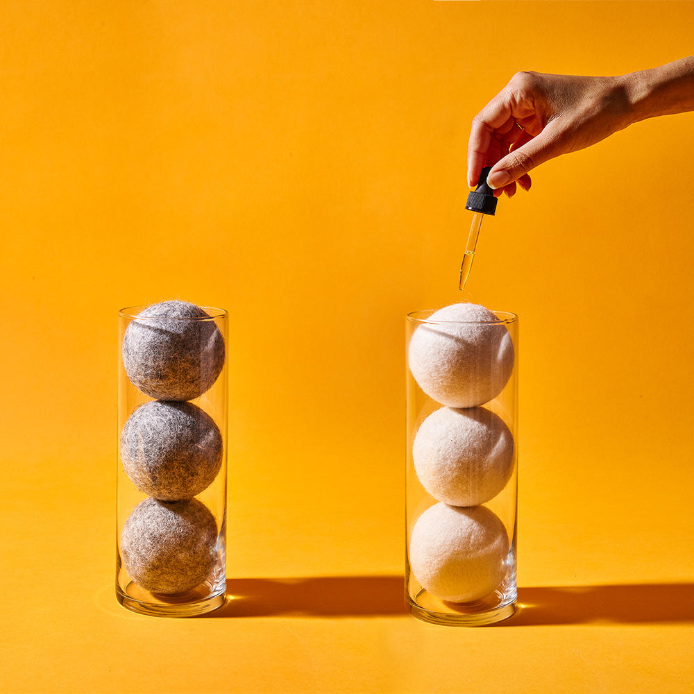 Six wool dryer balls in two glass display containers and a hand holding a dropper of essential oil