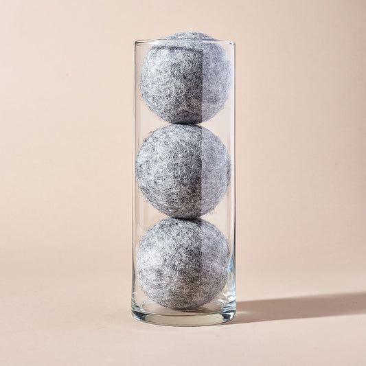 A stack of three grey wool dryer balls displayed in a clear tubular glass container with a beige backdrop