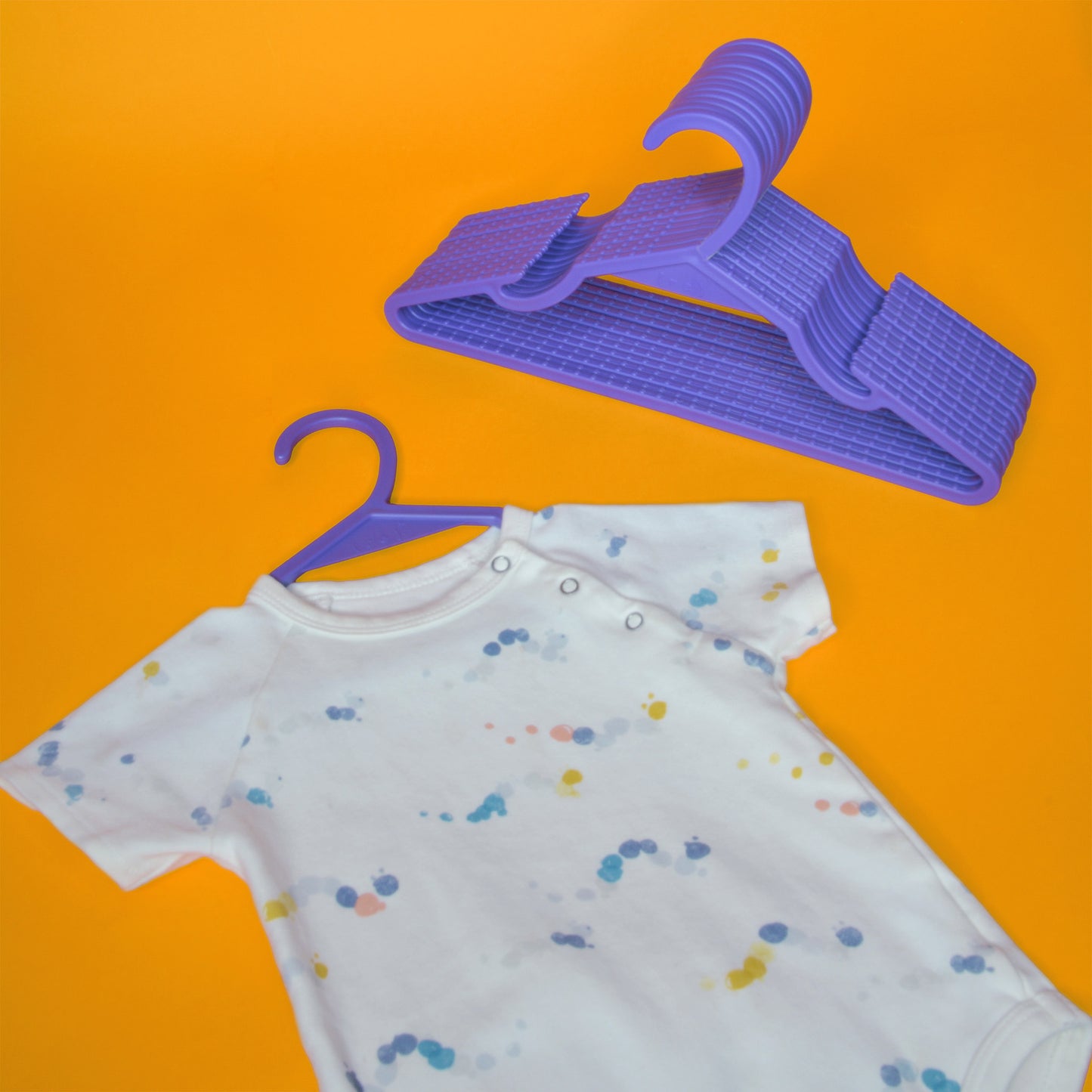 A colorful polka dot baby onesie with a set of bright purple kids hangers made from recycled ocean plastic