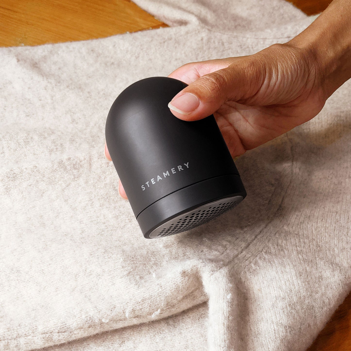 A tan hand holds a sleek dome shaped charcoal colored fabric shaver above a cream sweater; the word "Steamery" is written across the bottom of the device
