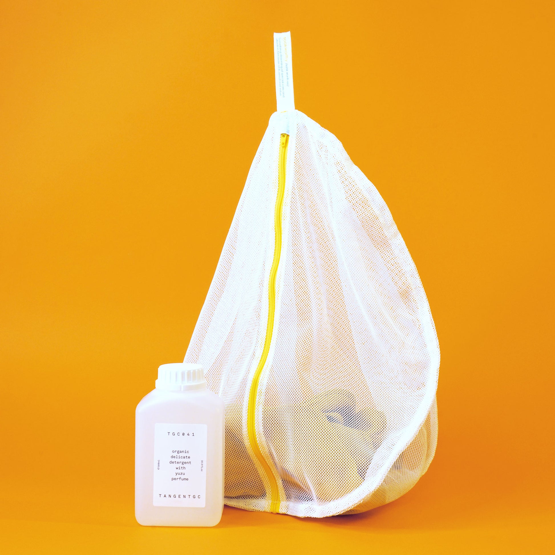 White mesh delicates bag by Soak with a yellow zipper; a piece of soft khaki clothing is visible inside and a bottle of delicate detergent is beside it