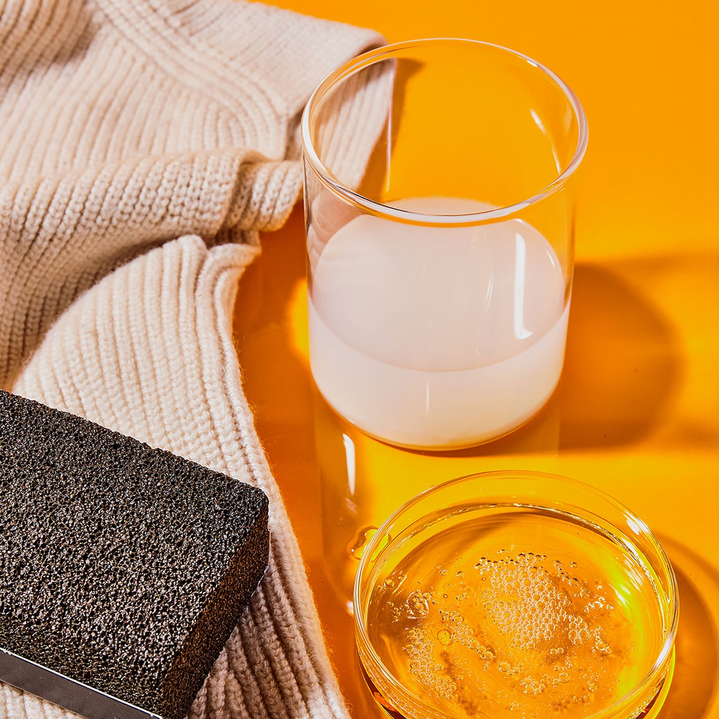 A black depilling "stone" displayed with liquid detergent in a clear petri dish, wool conditioner in a clear glass, and a beige knit sweater