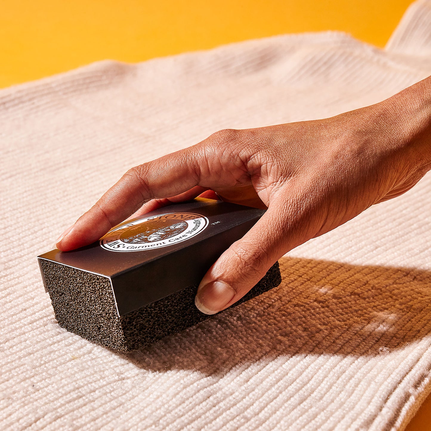 A hand brushes a rectangular pumice-like tool across a beige sweater to remove pilling