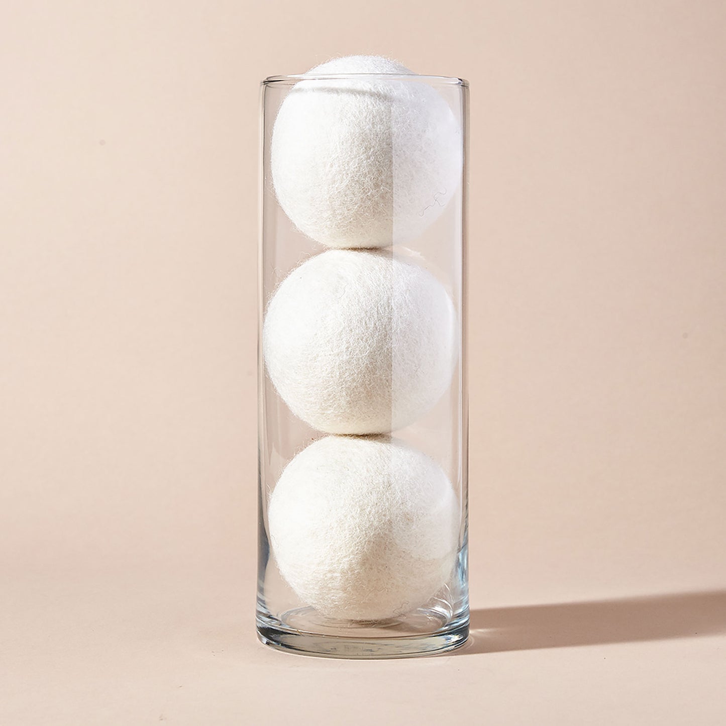 A stack of three white wool dryer balls displayed in a clear tubular glass container with a beige backdrop