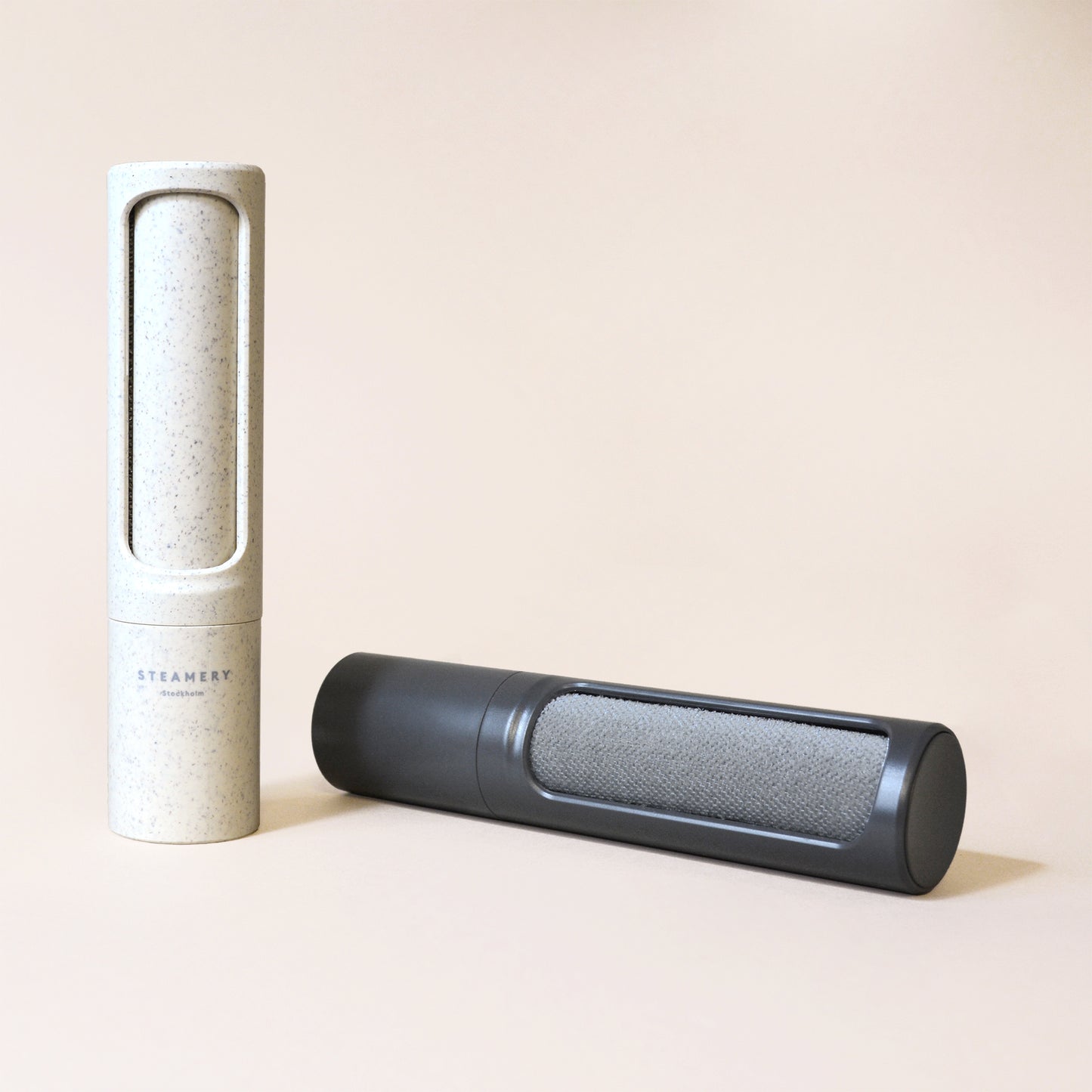 A self-standing speckled beige reusable lint roller next to a charcoal reusable lint roller with grey angled polyester bristles visible; both are on a beige backdrop