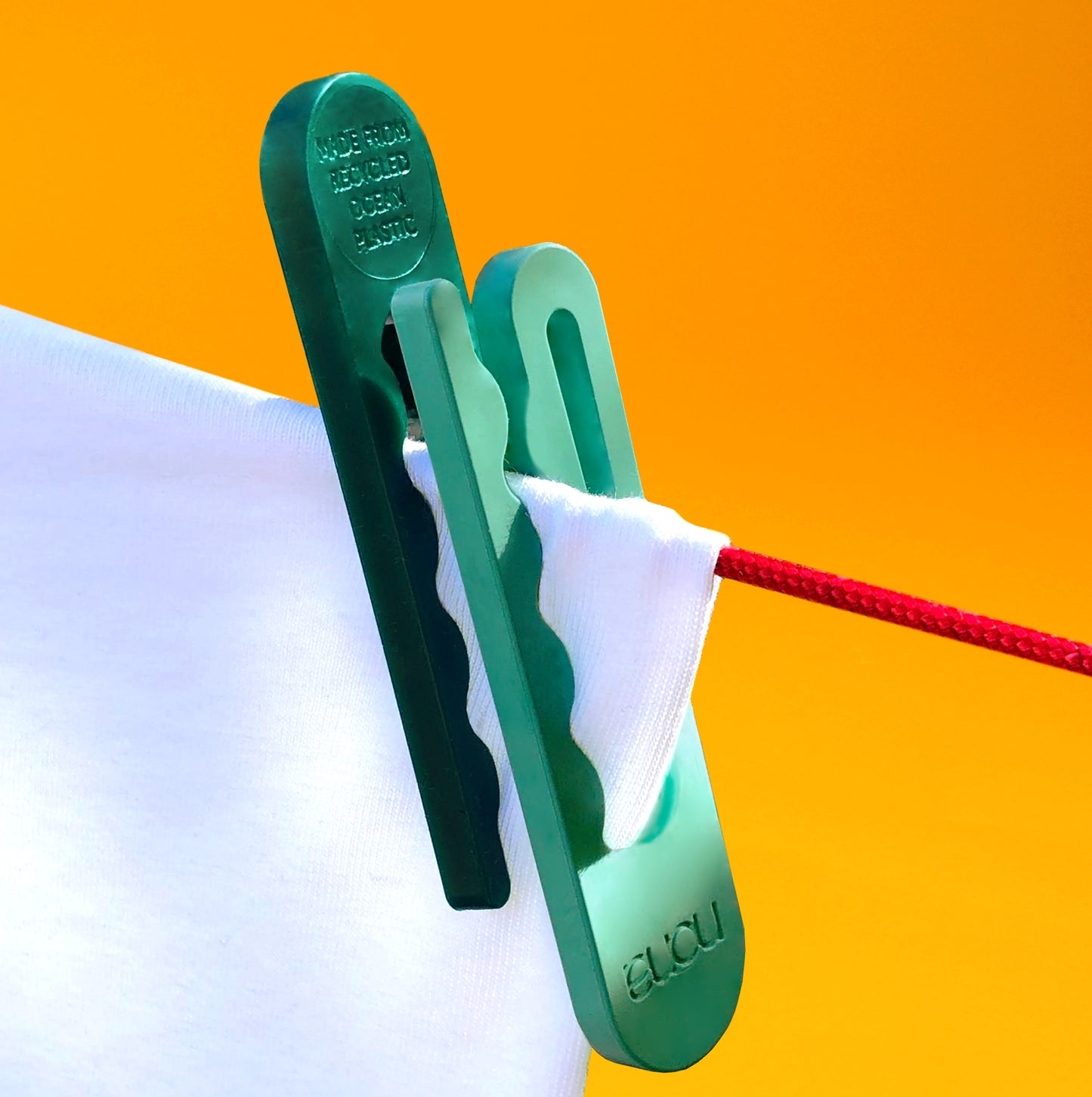 A light green recycled clothespin securing the corner of white fabric onto a red clothesline and a dark green clothespin pinned in the opposite direction