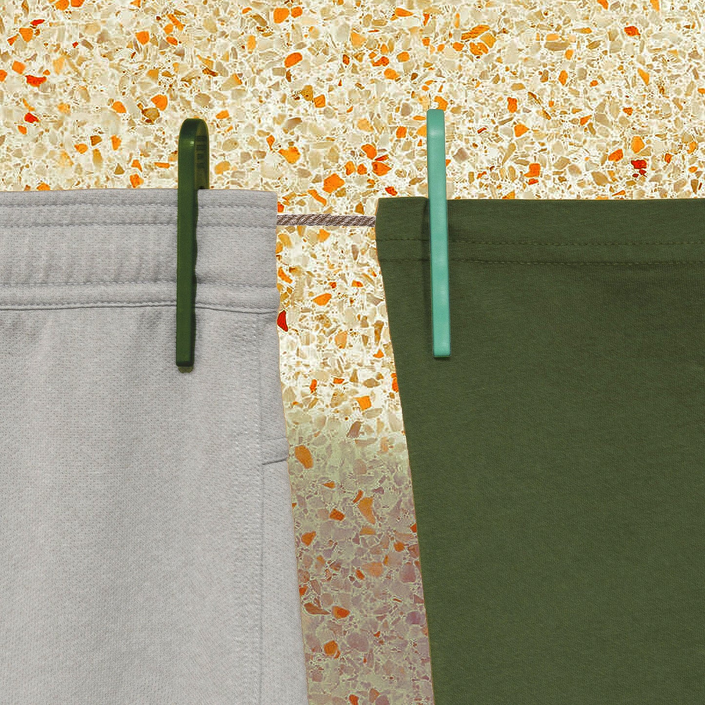 Grey shorts and a green t-shirt hang from a clothesline in front of a terrazzo wall; green ocean plastic clothespins hold them in place