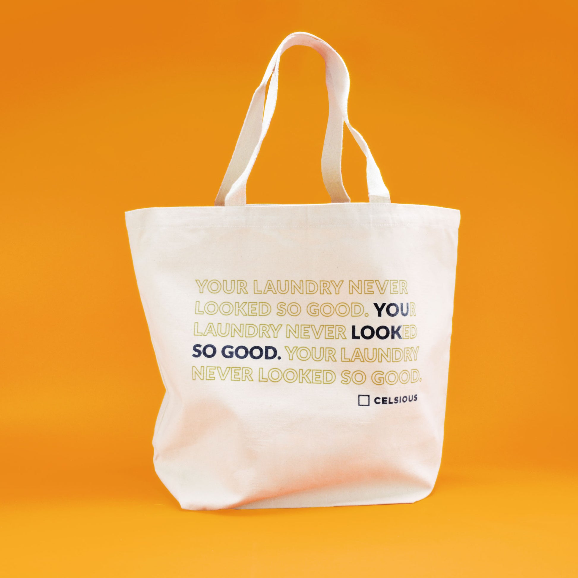 A beige Celsious tote bag with the words "Your laundry never looked so good" printed in navy and seaweed green, shot on an orange background